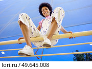 From below of delighted African American female looking at camera while sitting on railing with bent legs near glass building. Smiling black woman sitting on fence in city. Стоковое фото, агентство Ingram Publishing / Фотобанк Лори