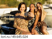 Smiling ethnic woman taking self portrait on cellphone with black girlfriend showing tongue and blonde making grimace on pier. Funny diverse women taking selfie on smartphone on seafront. Стоковое фото, агентство Ingram Publishing / Фотобанк Лори