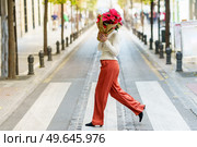Full body of unrecognizable female pedestrian in stylish outfit covering face with potted poinsettia plant with bright red leaves wrapped in paper while crossing road on crosswalk. Trendy woman with Euphorbia pulcherrima plant on zebra. Стоковое фото, агентство Ingram Publishing / Фотобанк Лори
