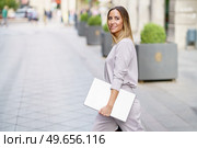 Side view of cheerful adult businesswoman with long blond hair in stylish outfit smiling, and looking away while strolling on paved street with laptop in hand before work. Happy female manager with laptop walking on street before meeting. Стоковое фото, агентство Ingram Publishing / Фотобанк Лори