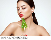 Beauty model girl eating Christmas tree shape lollipop.  Beautiful young woman with clean fresh skin. Girl beauty face care. Facial treatment. Cosmetology, beauty and spa. Red nails manicure. Стоковое фото, агентство Ingram Publishing / Фотобанк Лори