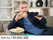 man changing tv channel with remote control while eating sandwich. Стоковое фото, агентство Ingram Publishing / Фотобанк Лори