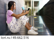 Full body young Asian woman in stylish clothes with cup of coffee to go siting on slope outside modern building and browsing smartphone in daytime. Asian female with coffee using cellphone on slope. Стоковое фото, агентство Ingram Publishing / Фотобанк Лори