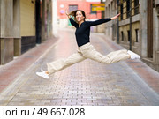 Side view of delighted young female with brown hair in casual clothes and sneakers smiling and looking at camera while jumping on narrow paved street. Excited young woman jumping on paved street and smiling. Стоковое фото, агентство Ingram Publishing / Фотобанк Лори