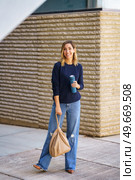 Full body cheerful woman in stylish outfit with bag and thermos standing on pavement and smiling near contemporary building on city street. Merry female against modern building. Стоковое фото, агентство Ingram Publishing / Фотобанк Лори