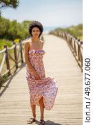 Full body young black woman with curly hair looking at camera, and waving skirt of long dress while standing on boardwalk leading to sea on sunny summer day. Ethnic tourist waving dress skirt. Стоковое фото, агентство Ingram Publishing / Фотобанк Лори