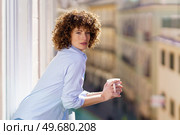 Adult female with brown curly hair leaning on railing of balcony while enjoying cup of hot beverage against blurred background. Curly haired woman drinking coffee on balcony. Стоковое фото, агентство Ingram Publishing / Фотобанк Лори