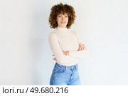 Confident adult curly haired female model wearing beige turtleneck and blue jeans standing with arms crossed against white background smiling at camera. Smiling woman with hands folded in studio. Стоковое фото, агентство Ingram Publishing / Фотобанк Лори