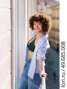 Adult positive female in unbuttoned shirt jeans and black bra standing on balcony against blurred background of town during weekend. Smiling curly haired woman in bra standing on balcony. Стоковое фото, агентство Ingram Publishing / Фотобанк Лори
