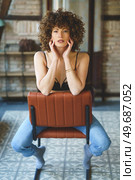 Full body of sensual young female, with afro hairstyle in black lingerie and blue jeans witting on leather chair and touching face while looking at camera against brick wall. Seductive curly haired woman in bra sitting on chair in room. Стоковое фото, агентство Ingram Publishing / Фотобанк Лори