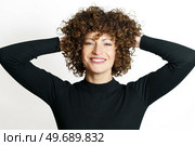 Positive young female model, with curly hair smiling and looking at camera while touching hands behind head against white background in unseen illuminating lights on shiny face. Happy woman with curly hair smiling with hands behind. Стоковое фото, агентство Ingram Publishing / Фотобанк Лори