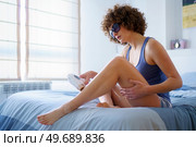 Adult woman in sleepwear and protective goggles, with curly hairstyle using IPL for knee hair removal session during skincare routine at home. Adult female using IPL for removing knee hair on bed with protective goggles. Стоковое фото, агентство Ingram Publishing / Фотобанк Лори
