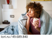 Thoughtful lady looking away and leaning on arm while drinking hot beverage and lounging on bed in morning. Wistful female with mug of tea wrapped in blanket. Стоковое фото, агентство Ingram Publishing / Фотобанк Лори