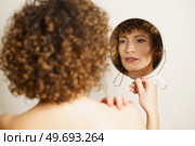 Beautiful young woman, with curly hair and makeup standing with back against camera while holding mirror in hand and looking away in reflection of self near gray background. Charming adult female with makeup looking reflection in mirror. Стоковое фото, агентство Ingram Publishing / Фотобанк Лори