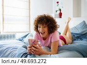 Delighted young woman with curly hair lying on comfortable bed while surfing social media via mobile phone in bedroom at home. Amazed woman using smartphone on bed at home. Стоковое фото, агентство Ingram Publishing / Фотобанк Лори