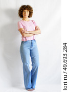 Full body of curly haired woman in casual clothes leaning on white wall while standing with hands crossed looking at camera and smiling. Full body of woman with curly hair standing against white wall crossing arms. Стоковое фото, агентство Ingram Publishing / Фотобанк Лори