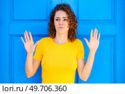 Scared young female in yellow t shirt with curly brown hair looking away with raised hands against blue background on city street. Frightened woman with raised hands on street. Стоковое фото, агентство Ingram Publishing / Фотобанк Лори
