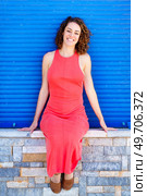 Full body of relaxing young female in red sundress sitting with hands on stonewall and smiling while looking at camera against blue shutters in daylight. Happy woman in red dress sitting on stone wall. Стоковое фото, агентство Ingram Publishing / Фотобанк Лори