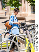 Side view of focused middle aged female in trendy outfit with handbag standing and using kick sharing service for rent electronic scooter. Mature woman renting scooter on street. Стоковое фото, агентство Ingram Publishing / Фотобанк Лори