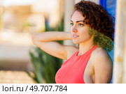 Side view of attractive young female in red sundress looking away while standing with hand on head on blurred background of street with blue shutters. Charming young woman standing with hand on head. Стоковое фото, агентство Ingram Publishing / Фотобанк Лори