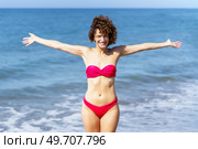 Cheerful young curly ginger haired female in pink bikini with outstretched hands and looking at camera, while standing near foamy rippling seawater against waving sea and blue sky. Happy woman in pink bikini standing near sea. Стоковое фото, агентство Ingram Publishing / Фотобанк Лори
