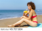 Side view of redhead woman in bikini sitting on beach of ocean in sunlight and drinking orange juice with straw from glass cup. Lounging lady with juice on coastline. Стоковое фото, агентство Ingram Publishing / Фотобанк Лори