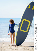 Full body view of anonymous young hairy female surfer in blue swimsuit standing with paddleboard on sandy beach near sea on sunny daylight. Unrecognizable surfer with paddleboard on sandy beach. Стоковое фото, агентство Ingram Publishing / Фотобанк Лори