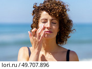 Young female with curly hair applying sunblock cream on face while standing against blurred sea on sunny day. Sensual woman applying sunscreen on face. Стоковое фото, агентство Ingram Publishing / Фотобанк Лори