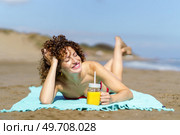 Cheerful young woman with curly hair smiling and closing eyes in sunlight while suntanning and drinking juice on sandy beach. Dreamy woman lying on sandy beach. Стоковое фото, агентство Ingram Publishing / Фотобанк Лори