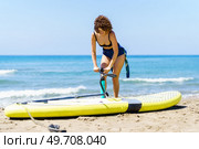 Focused young female with curly red hair in swimwear standing near paddleboard and pumping air, with handpump while looking down against blurred waving sea and blue sky. Slim woman standing with handpump near SUP board. Стоковое фото, агентство Ingram Publishing / Фотобанк Лори