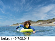 Young curly haired woman in wetsuit lying on standup SUP board paddling with hands in sea against blue sky on sunny summer day. Fit woman on paddleboard rowing with hands in ocean. Стоковое фото, агентство Ingram Publishing / Фотобанк Лори