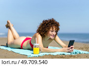 Happy woman with ginger curly hair in red bikini lying on coast of ocean with refreshing drink and texting on smartphone. Laughing woman using phone on coastline. Стоковое фото, агентство Ingram Publishing / Фотобанк Лори