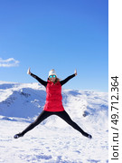Full body of excited young female in vest beanie cap polarized sunglasses looking at camera, while leaping in air with widened legs stretched arms against snowy mountains and blue sky. Carefree young woman jumping with raised arms in snowy terrain. Стоковое фото, агентство Ingram Publishing / Фотобанк Лори