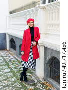 Full body of smiling female in trendy red coat and beret looking at camera while standing near white fencing in city. Cheerful woman in stylish outfit near fence. Стоковое фото, агентство Ingram Publishing / Фотобанк Лори