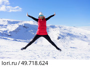 Full body of excited young female in warm clothes cap, polarized sunglasses looking at camera while jumping in air with widened legs stretched arms against snowy mountains and blue sky. Cheerful woman in warm clothes jumping in air on snow terrain. Стоковое фото, агентство Ingram Publishing / Фотобанк Лори