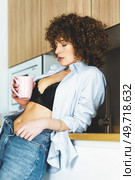 Curly haired woman in black bra unbuttoned shirt and unzipped jeans leaning on counter while enjoying hot beverage. Woman with unbuttoned shirt drinking coffee in kitchen. Стоковое фото, агентство Ingram Publishing / Фотобанк Лори