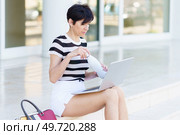 Concentrated adult female freelancer in striped t shirt sitting on stairs on street and looking at screen of laptop while working remotely and opening bottle. Focused woman with laptop opening thermos on stairs. Стоковое фото, агентство Ingram Publishing / Фотобанк Лори