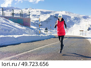 Full body of young female in red vest beanie cap, polarized sunglasses looking at camera while jogging on asphalt road near snow against snowy mountain and blue sky in daylight. Young woman running on road near snow and warming up. Стоковое фото, агентство Ingram Publishing / Фотобанк Лори