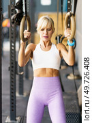 Determined fit female doing exercise with double circle ring and adjustable straps during fitness workout at modern gym club against blurred background looking down. Strong young woman exercising with double circle ring and adjustable straps. Стоковое фото, агентство Ingram Publishing / Фотобанк Лори