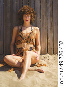 Full body of young female wearing swimsuit, and crochet beach dress sitting on warm sand against woodenit fence looking at camera while resting during summer weekend. Young lady in knitted beach cover up sitting on sandy shore. Стоковое фото, агентство Ingram Publishing / Фотобанк Лори