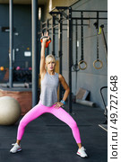 Focused adult female in sportswear looking at camera, while standing with legs apart lifting kettle ball over head and with hand on waist doing exercise in gym with equipment in daylight. Adult woman doing exercises with kettlebell in gym. Стоковое фото, агентство Ingram Publishing / Фотобанк Лори