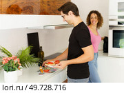 Side view of happy adult male looking down while standing with smiling conversing female at counter, with wine bottle flower pot and preparing healthy salad in kitchen at home in daylight. Happy adult man standing near cooking range with woman and arranging salad in kitchen. Стоковое фото, агентство Ingram Publishing / Фотобанк Лори