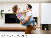 Side view of smiling young couple embracing while having romantic moment in modern apartment against LED TV and looking at each other happily. Cheerful young couple hugging at home. Стоковое фото, агентство Ingram Publishing / Фотобанк Лори