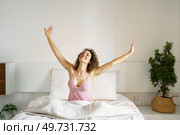 Smiling adult female with curly hair and with eyes closed, while sitting on cozy bed in daylight and stretching arms sideways with open palms in bedroom with green plant. Cheerful woman sitting on bed in sleepwear. Стоковое фото, агентство Ingram Publishing / Фотобанк Лори