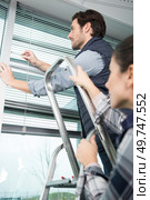 handyman repairing window blind his partner holding the stepladder. Стоковое фото, агентство Ingram Publishing / Фотобанк Лори