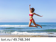 Full body side view of excited barefoot young female, in bikini with outstretched arms leaping with legs in air while looking away in daylight against rippling foamy seawater. Slim woman in bikini jumping on sandy beach. Стоковое фото, агентство Ingram Publishing / Фотобанк Лори