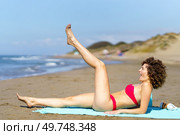 Side view of young woman with curly hair in pink bikini raising leg while having fun on sandy shoreline. Young woman in bikini chilling on sandy beach. Стоковое фото, агентство Ingram Publishing / Фотобанк Лори