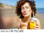 Young female with curly hair and closed eyes blowing kiss and holding jar of orange juice while taking selfie on beach during summer vacation. Playful woman pouting lips while having juice on beach. Стоковое фото, агентство Ingram Publishing / Фотобанк Лори