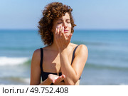 Sensual woman in bikini standing near waving ocean while applying sunblock cream on face during summer vacation. Young female smearing cream on face protecting from sunburn. Стоковое фото, агентство Ingram Publishing / Фотобанк Лори