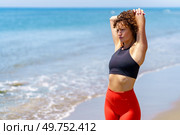 Thoughtful young curly haired sportswoman in black top and red leggings stretching arm during warm up on beach while looking away. Pensive woman stretching on seashore. Стоковое фото, агентство Ingram Publishing / Фотобанк Лори